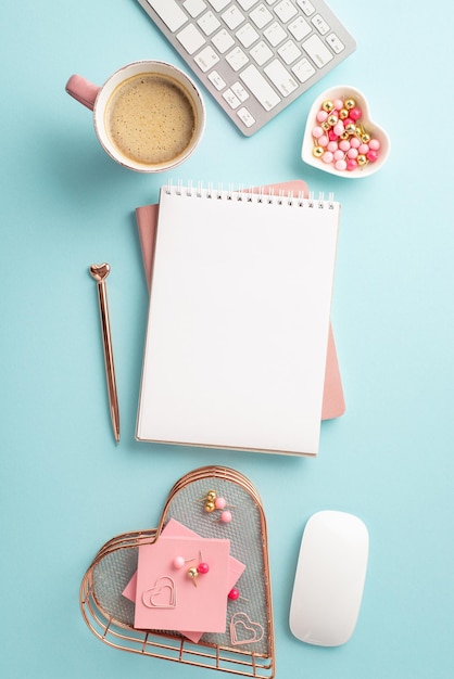 Vista superior foto vertical de blocos de anotações caneta teclado computador rato porta-papéis em forma de coração com pushpins papel de nota pegajoso e xícara de café em fundo azul pastel isolado com espaço vazio