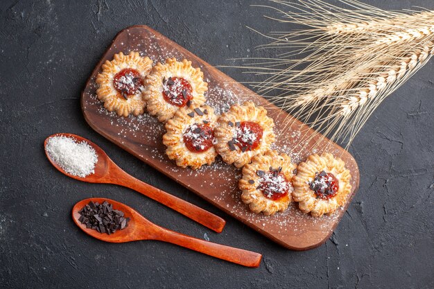 Vista superior de galletas con mermelada sobre tablero de madera de coco en polvo y chocolate en polvo en cucharas de madera espigas de trigo sobre fondo oscuro