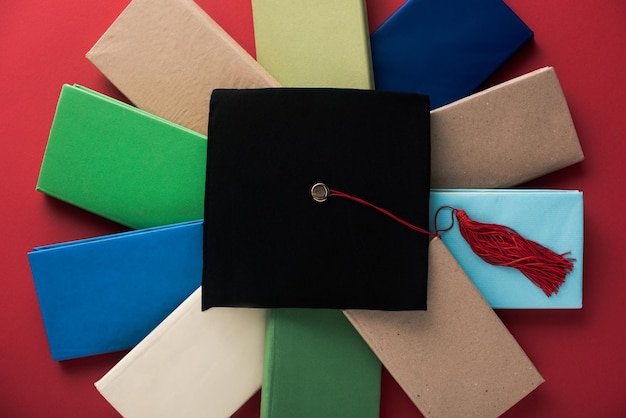 Vista superior de la gorra de graduación negra con borla en libros sobre fondo rojo.