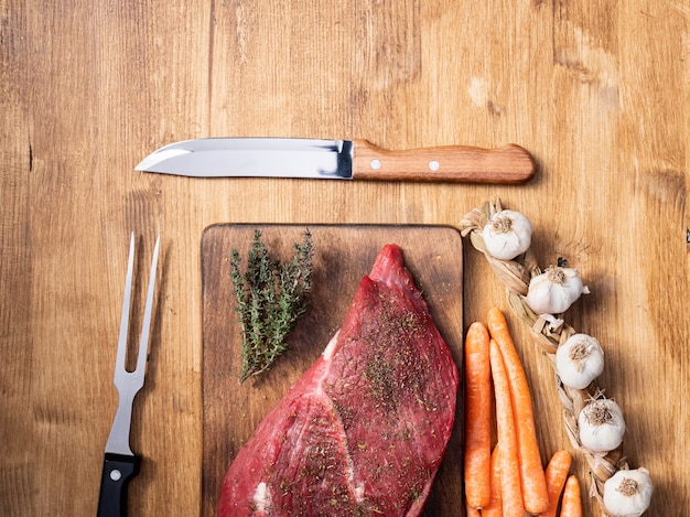 Foto vista superior de un gran trozo de carne roja junto a ajo, zanahorias y cuchillo de chef en el centro del marco. preparación de alimentos. proteína natural.