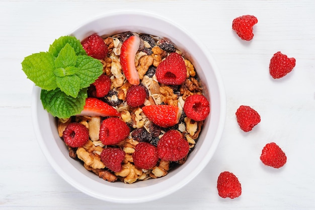 Vista superior de una granola orgánica casera con nueces de avena, frambuesas y fresas en un tazón blanco sobre una tabla vintage de madera