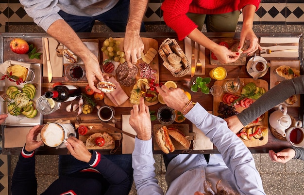 Vista superior del grupo de amigos sin rostro comiendo deliciosos bocadillos mientras se reúnen en una mesa de madera con bebidas en una cocina ligera