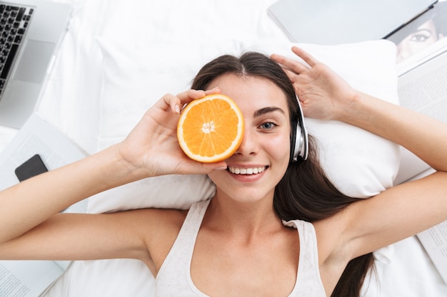 Vista superior de una hermosa joven escuchando música con auriculares mientras está acostado en la cama, sosteniendo una rodaja de naranja