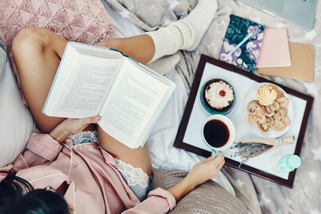 Vista superior de la hermosa joven en pijama leyendo un libro y disfrutando del café de la mañana mientras descansa en la cama en casa