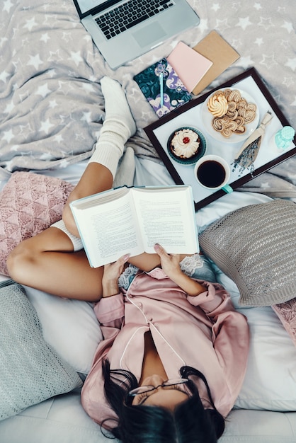Vista superior de la hermosa joven en pijama leyendo un libro y disfrutando del café de la mañana mientras descansa en la cama en casa