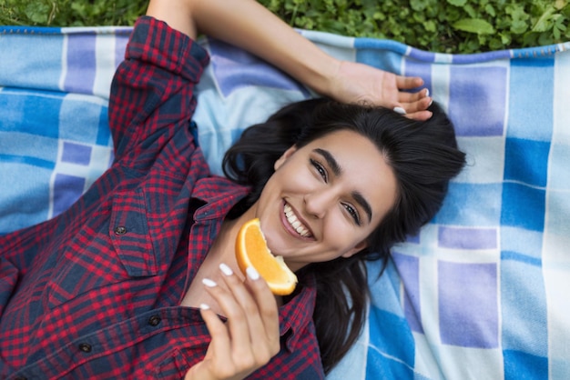 Vista superior de hermosa mujer sonriente morena bonita sosteniendo jugosa naranja deliciosa en el parque verde de verano Sonrisa saludable femenina joven disfrutando y sosteniendo fruta naranja para un estilo de vida vegetariano