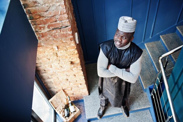 Vista superior del hombre africano en ropa tradicional negra con gorra posada en el restaurante en las escaleras