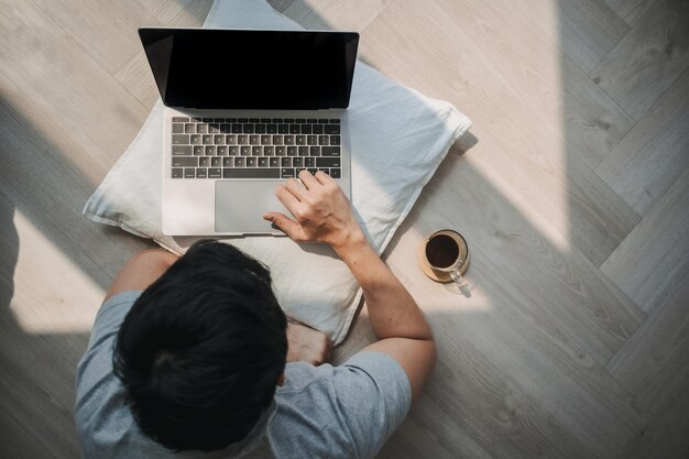 Vista superior de un hombre asiático usando una computadora tendida en el suelo mientras trabaja en una computadora portátil con café y escritura