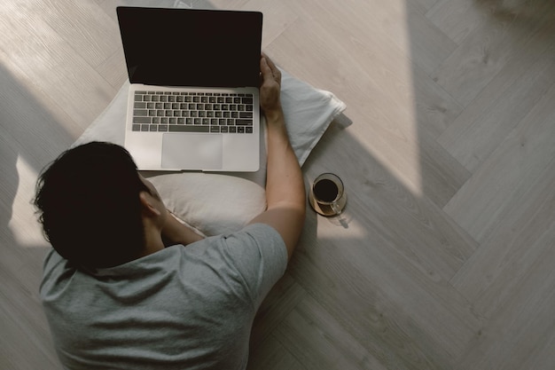 Foto vista superior de un hombre asiático usando una computadora tendida en el suelo mientras trabaja en una computadora portátil con café y escritura