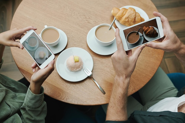 Vista superior del hombre irreconocible que toma una foto de café negro y croissant en la mesa en el café blogger