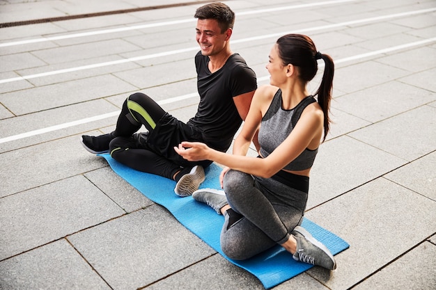 Vista superior del hombre y la mujer atléticos sonrientes que desarrollan la flexibilidad corporal mientras hacen ejercicio en la colchoneta en el centro de la ciudad