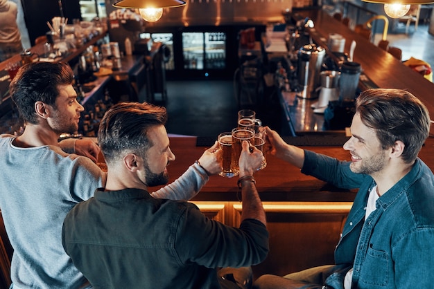 Vista superior de hombres jóvenes felices en ropa casual brindando con cerveza y sonriendo mientras está sentado en el pub