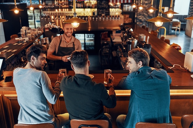 Vista superior de hombres jóvenes relajados en ropa casual bebiendo cerveza mientras están sentados en el pub