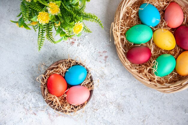 Vista superior de huevos de pascua de colores dentro de la placa y la canasta con flores sobre una superficie blanca