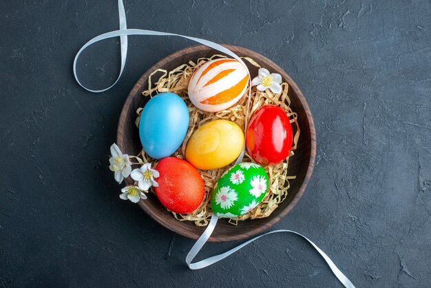 Vista superior de los huevos de pascua de colores dentro de la placa con paja sobre una superficie oscura