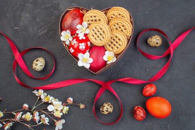 Vista superior de huevos de pascua de colores con galletas dentro de una caja en forma de corazón sobre una superficie oscura