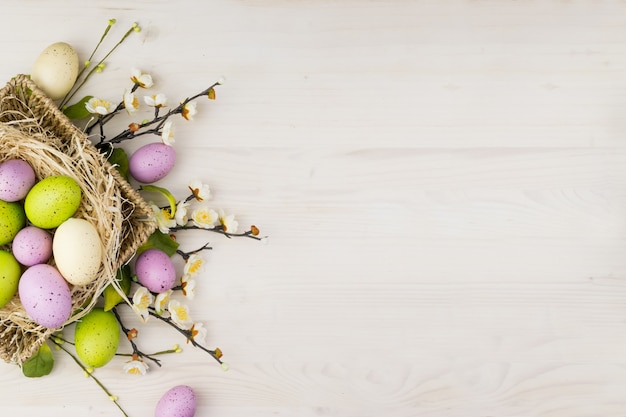 La vista superior de los huevos de Pascua coloridos en cesta y la primavera florece en un fondo de madera claro con el espacio del mensaje.