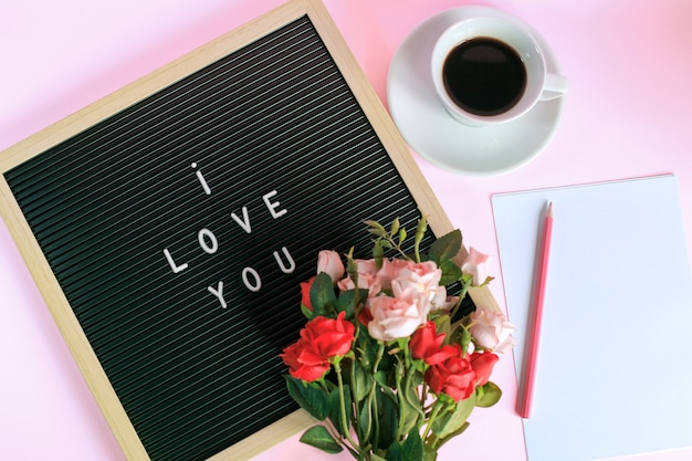 Vista superior de I LOVE YOU en tablero de letras con taza de café, rosas y lápiz sobre papel en blanco aislado sobre fondo rosa