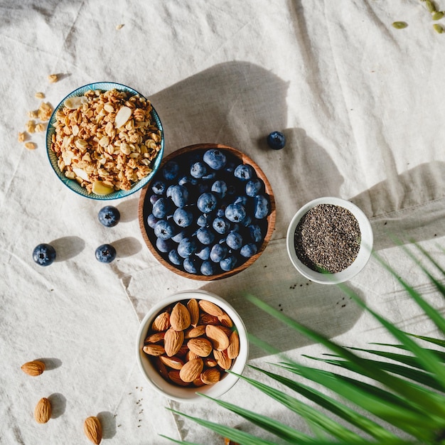 Vista superior de los ingredientes del desayuno como cereales, almendras, arándanos y semillas de chía en un paño de lino decorado con una hoja de palma