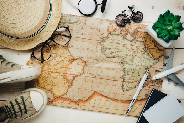 Vista superior itens de viagem essenciais. O saco da calculadora, caderno, árvore, mapa, passaporte, avião, auscultadores, música, óculos, branca, madeira, fundo.