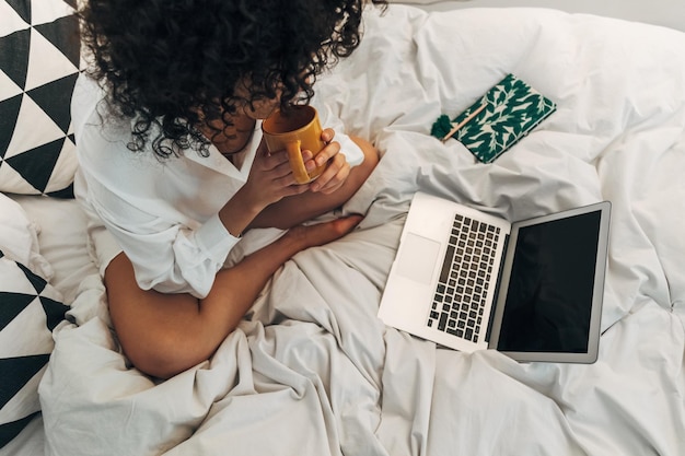 Vista superior de una joven afroamericana que trabaja usando una laptop en casa sentada en la cama bebiendo té Tecnología