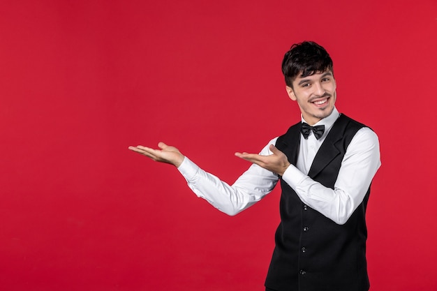 Vista superior del joven camarero masculino confiado sonriente en uniforme con pajarita y apuntando hacia arriba en el lado derecho con ambas manos