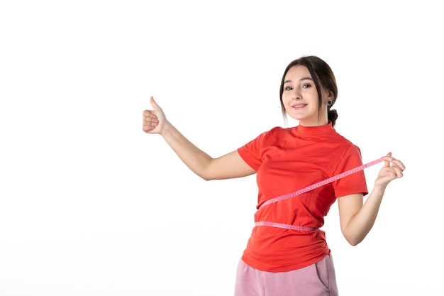 Vista superior de una joven confiada recogiendo su cabello vestida con una blusa redorange y sosteniendo un medidor midiendo su cintura haciendo un gesto de ok sobre fondo blanco