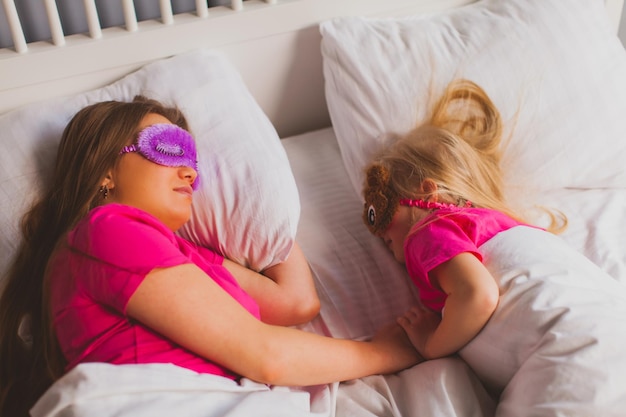 Vista superior de la joven madre y su pequeña hija duermen uno frente al otro Se acuestan en la cama con divertidas máscaras para dormir