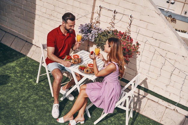Vista superior de la joven pareja en ropa casual desayunando y sonriendo mientras está sentado en el patio de la azotea al aire libre