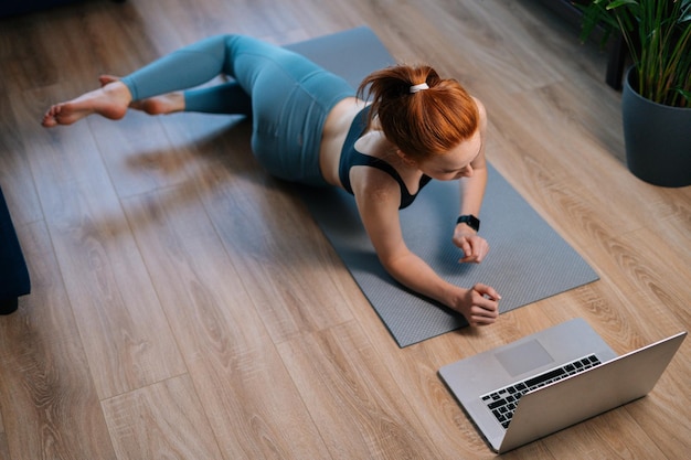 Vista superior de una joven pelirroja en forma haciendo ejercicio, haciendo ejercicio de estiramiento en una alfombra de yoga mientras ve videos de fitness en línea en una laptop. Concepto de entrenamiento deportivo dama pelirroja durante la cuarentena.