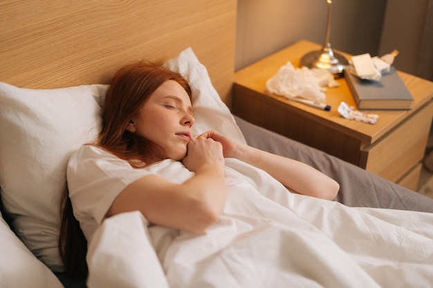 Vista superior de una joven pelirroja tranquila y enferma con ropa de noche suave durmiendo en una cama cómoda acostada sobre una almohada suave