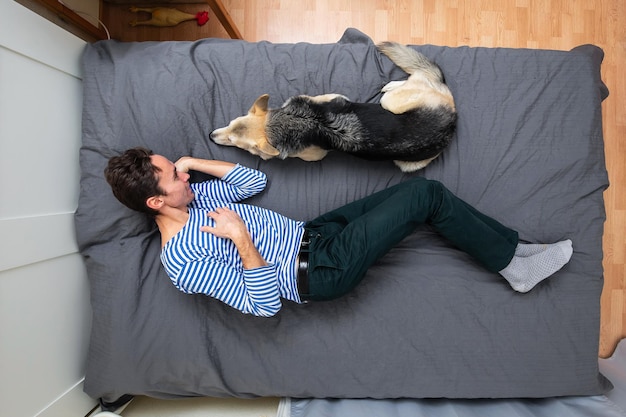 Vista superior de un joven relajado con camisa a rayas acostado con un perro mestizo en la cama con una sábana gris