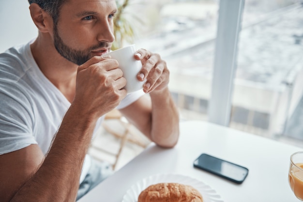 Vista superior del joven relajado disfrutando de un café caliente mientras está sentado en la cocina