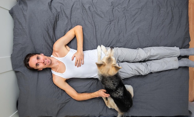 Vista superior de un joven sonriente con ropa de casa acostado con perros mestizos en la cama con una sábana gris