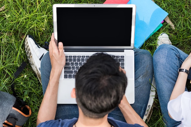 Vista superior jovens estudantes sentados na grama com laptop e livro consultando aprendendo juntos