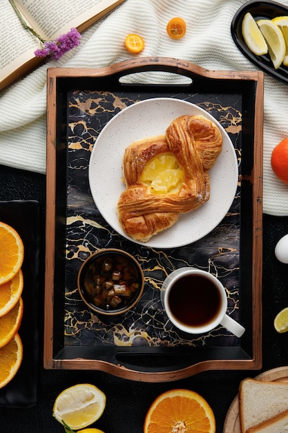 Vista superior del juego de desayuno con croissant en plato y mermelada en tazón y taza de té en bandeja con rodajas de naranja y limón con libro abierto y florecita en libro sobre tela sobre fondo negro