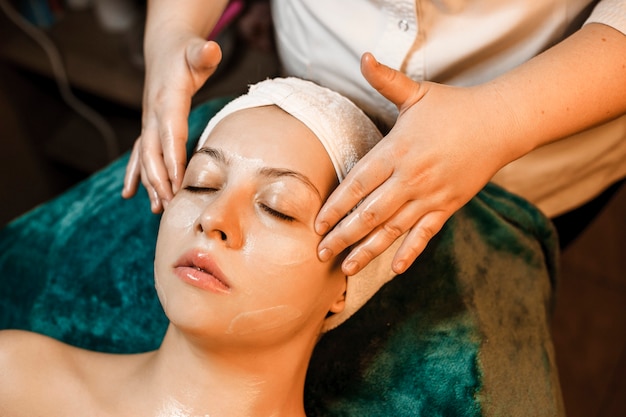 Vista superior de una linda mujer que recibe un masaje facial por parte de una cosmetóloga mientras tiene una máscara transparente en su rostro apoyado en una cama con los ojos cerrados.