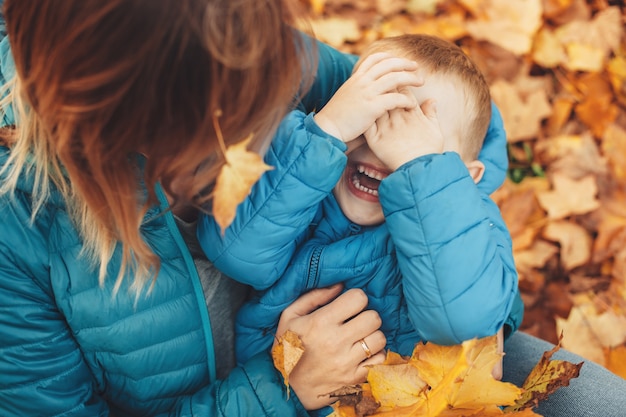 Vista superior de un lindo niño riendo mientras se apoya en las piernas de su madre ocultando su rostro con las manos.