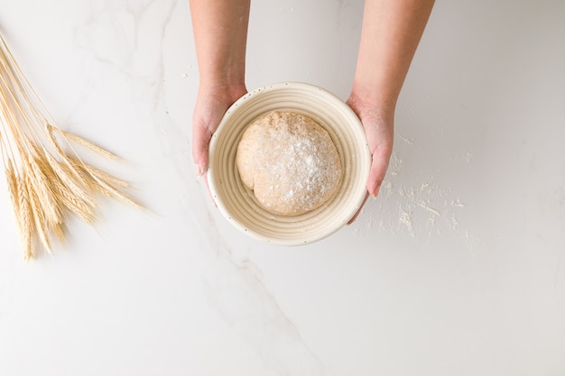 Foto vista superior de la mano femenina sosteniendo una masa de pan en reposo en un bol de pan en una mesa de mármol con trigo y harina con espacio para texto