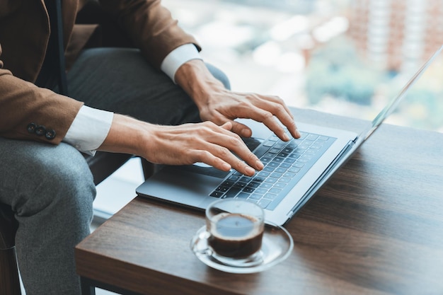 Vista superior de la mano de un hombre de negocios inteligente que busca, escribe y analiza datos utilizando una computadora portátil Imagen en primer plano de las manos del gerente trabajando con la computadora portatil en un escritorio de madera con una taza de café rodeada de naturaleza Ornamentada