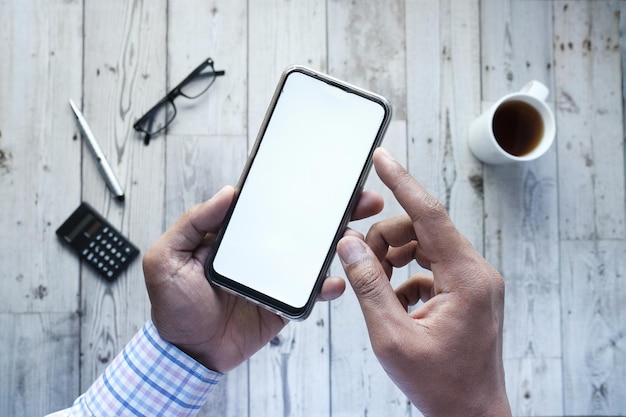 Vista superior de la mano del hombre que sostiene el teléfono inteligente con pantalla vacía en el escritorio de la oficina