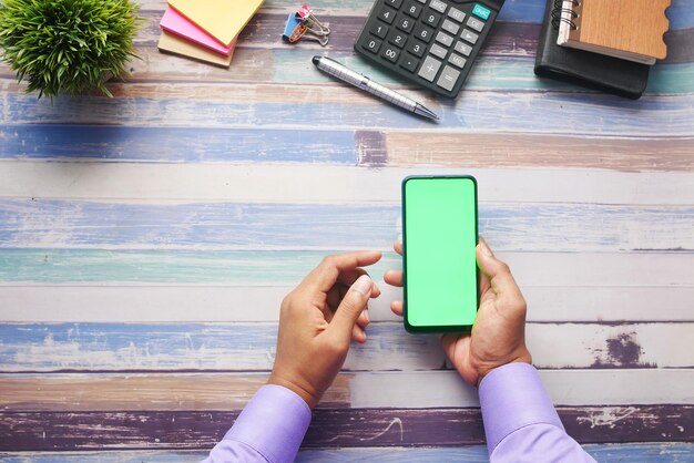 Vista superior de la mano del hombre sosteniendo un teléfono inteligente con pantalla vacía en el escritorio de la oficina