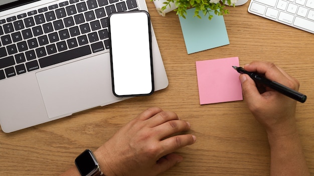 Vista superior de la mano masculina con lápiz escribiendo en el bloc de notas mientras trabaja con la computadora portátil y el teléfono inteligente en la mesa de madera