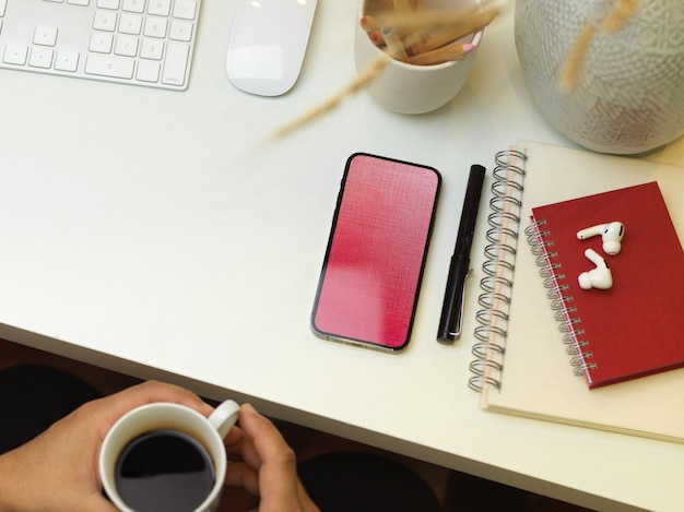 Vista superior de la mano masculina sosteniendo la taza de café mientras está sentado en el espacio de trabajo con teléfono inteligente, artículos de papelería y suministros