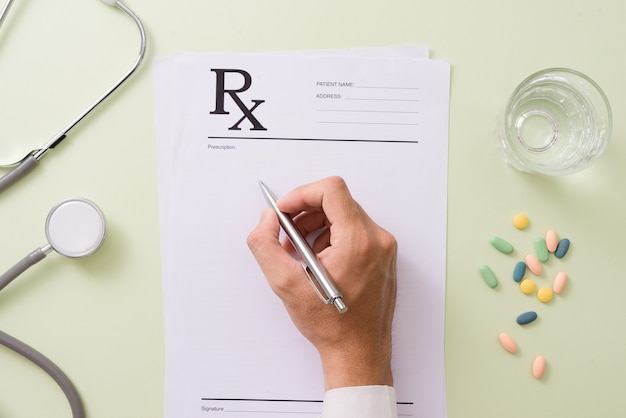 Vista superior de la mano de un médico escribiendo una receta. Un frasco de pastillas y un estetoscopio también están en el escritorio.