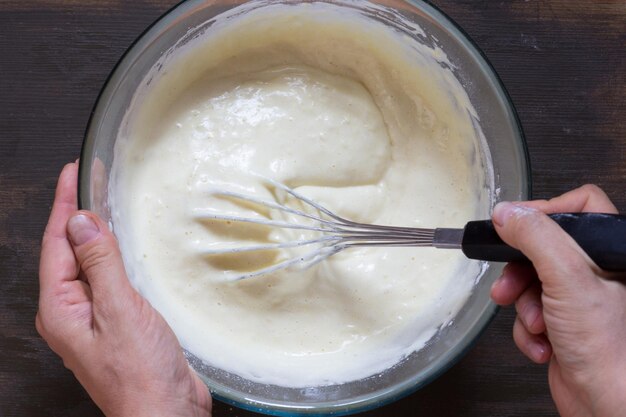 Vista superior de la mano de la mujer mezclando con ingredientes secos batidores y masa en el fondo de madera