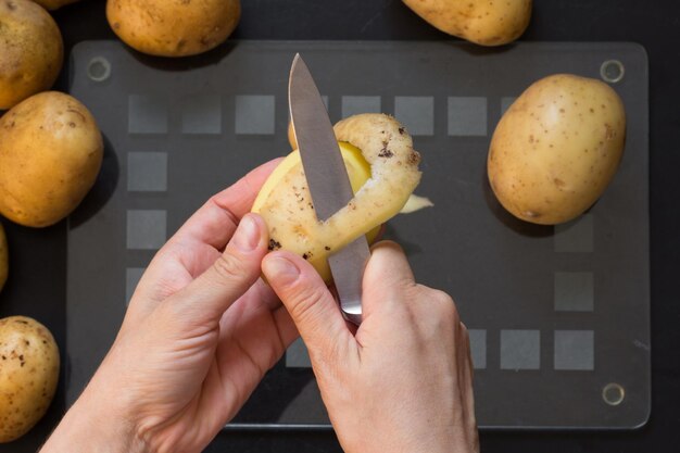 Vista superior de la mano de la mujer pelando patatas crudas con un cuchillo en el fondo negro