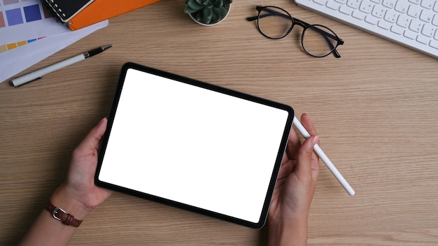 Vista superior de la mano de la mujer que sostiene la tableta digital con pantalla en blanco y lápiz óptico en la mesa de madera.