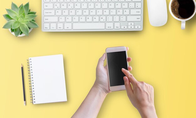 Vista superior con mano de mujer usando teléfono móvil con pantalla vacía sobre fondo de mesa amarillo