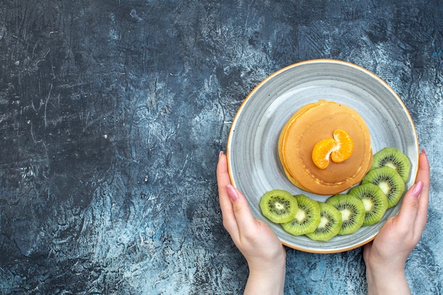 Vista superior de la mano sosteniendo esponjosos panqueques estilo americano hechos con yogur natural servido con kiwi y mandarina en un plato en el lado izquierdo sobre fondo de hielo con espacio libre